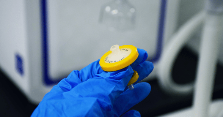 Gloved hand holding a yellow syringe filter in a lab setting.